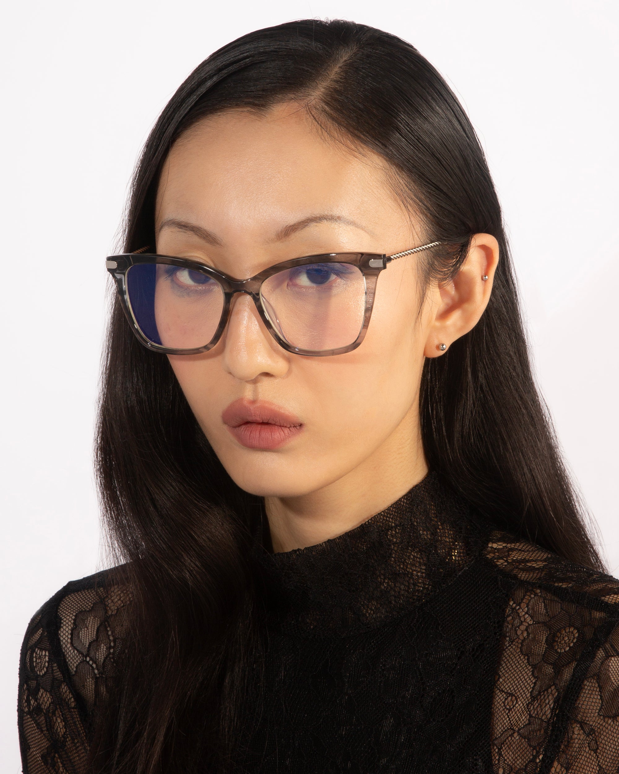 A person with long, straight black hair is wearing the Paris Two clear-rimmed glasses with a blue light filter from For Art's Sake®. They have a serious expression and are looking directly at the camera, dressed in a black lace top. The background is plain white.