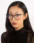 A person with long, straight black hair is wearing the Paris Two clear-rimmed glasses with a blue light filter from For Art's Sake®. They have a serious expression and are looking directly at the camera, dressed in a black lace top. The background is plain white.