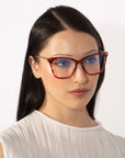 A woman with long, straight black hair and minimal makeup is wearing the Paris Two red-framed eyeglasses with a blue light filter from For Art's Sake®. She is dressed in a sleeveless, pleated white top against a plain white background.