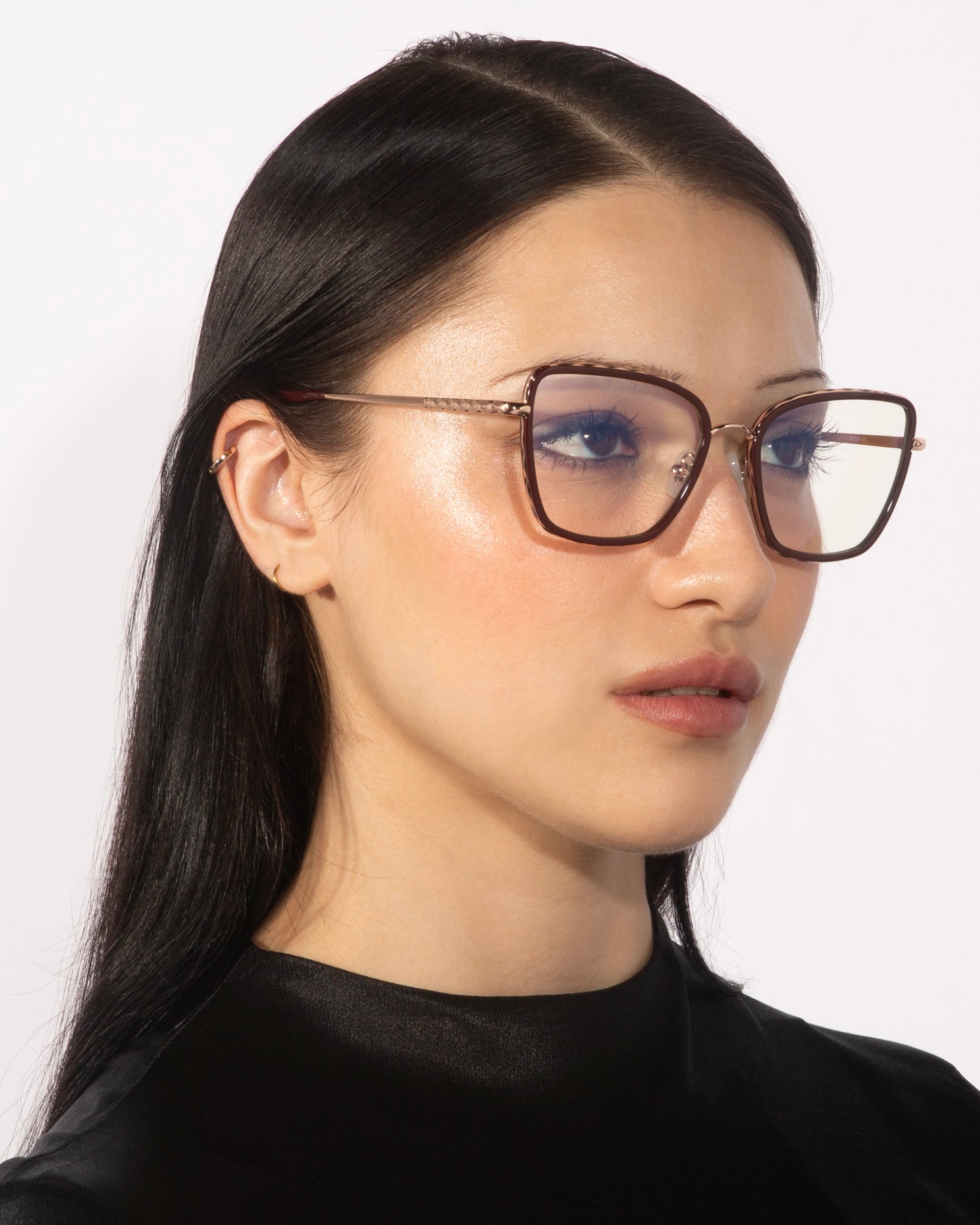 A person with long black hair and glowing skin is wearing oversized, square-shaped glasses named "Lace" by For Art's Sake®, featuring 18-karat gold-plated frames. They are dressed in a black top and are gazing slightly to the side against a white background.