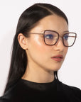 A person with long black hair and glowing skin is wearing oversized, square-shaped glasses named "Lace" by For Art's Sake®, featuring 18-karat gold-plated frames. They are dressed in a black top and are gazing slightly to the side against a white background.