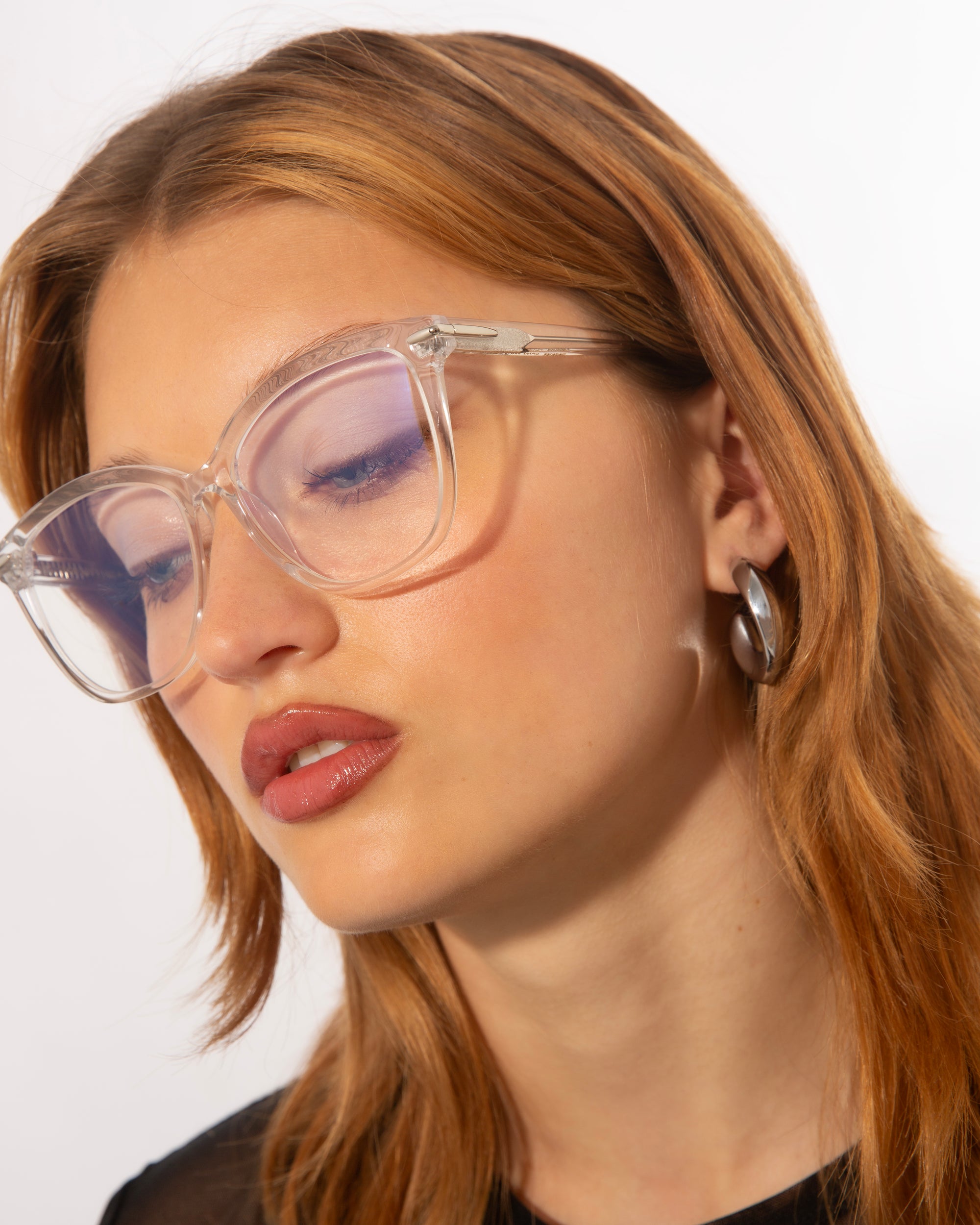 A person with light brown hair is wearing large, transparent Cadenza optical glasses by For Art's Sake® and hoop earrings. They have a neutral expression and are looking downward slightly. The background is plain white, highlighting their features.