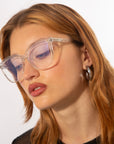 A person with light brown hair is wearing large, transparent Cadenza optical glasses by For Art's Sake® and hoop earrings. They have a neutral expression and are looking downward slightly. The background is plain white, highlighting their features.