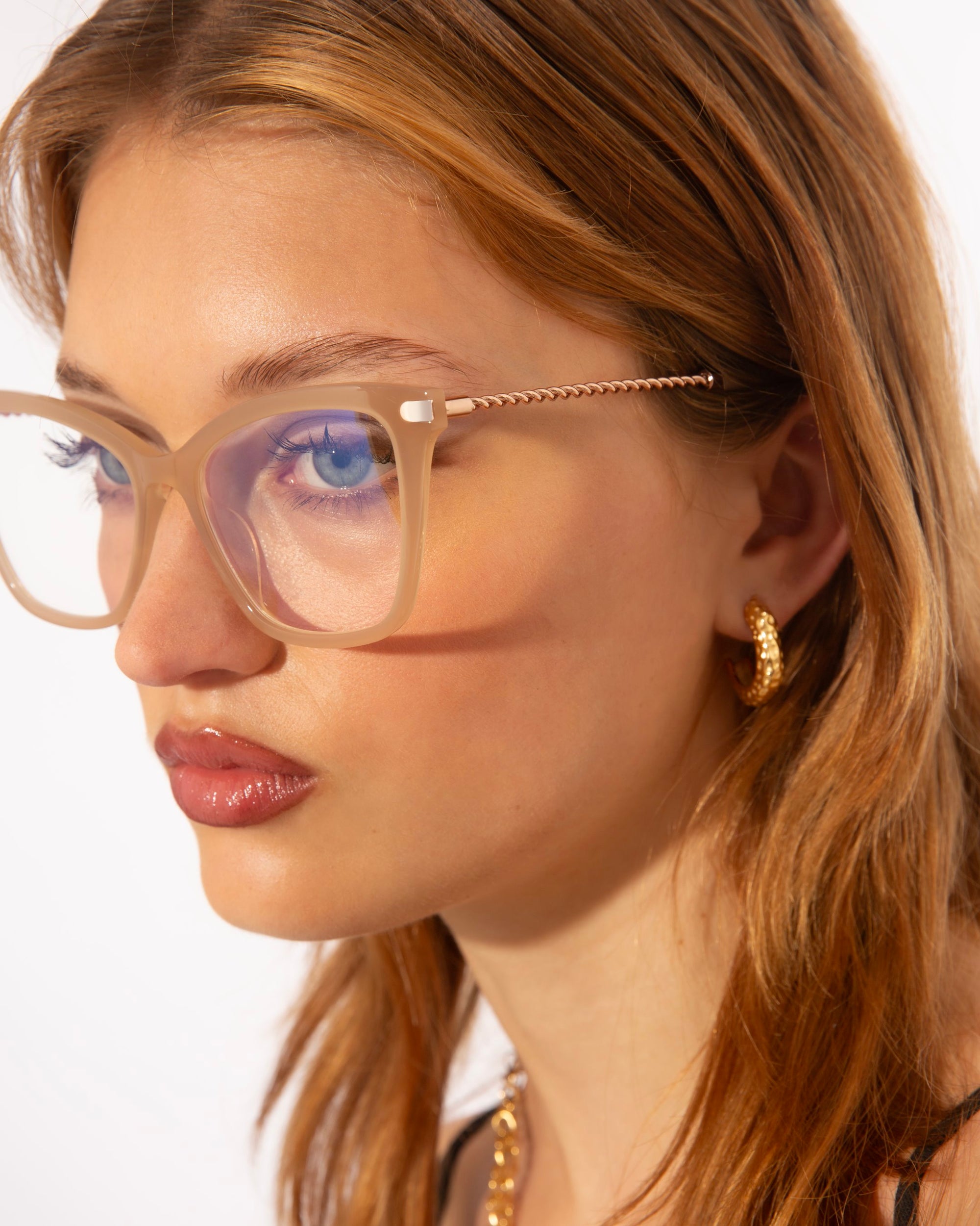 A close-up of a young person with long, light brown hair, wearing large, translucent beige acetate Cadenza glasses in a cat-eye silhouette from For Art&#39;s Sake®. They have a neutral expression and wear a small, twisted gold hoop earring. The background is white, highlighting their features and accessories.