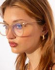 A close-up of a young person with long, light brown hair, wearing large, translucent beige acetate Cadenza glasses in a cat-eye silhouette from For Art's Sake®. They have a neutral expression and wear a small, twisted gold hoop earring. The background is white, highlighting their features and accessories.