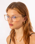 A young woman with long, light brown hair wears Cadenza rectangular optical glasses by For Art's Sake®, a gold chain necklace with pearl accents, and gold hoop earrings. She has a serious facial expression and is posed against a plain white background.