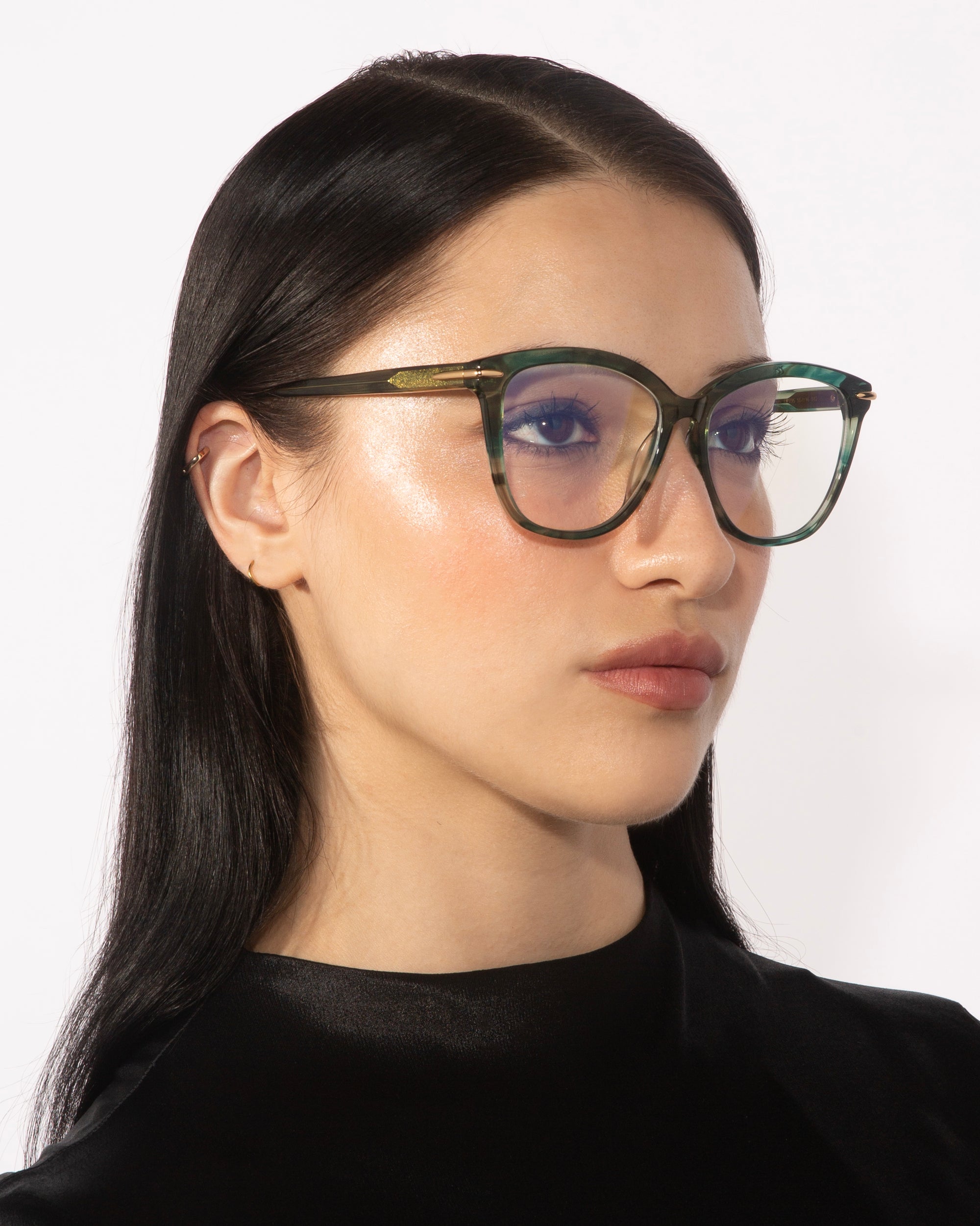 A woman with long black hair is wearing the stylish Cadenza cat-eye silhouette glasses in chic green and clear acetate from For Art's Sake®. She gazes to the side with a neutral expression, dressed in a sleek black top against a plain white background.