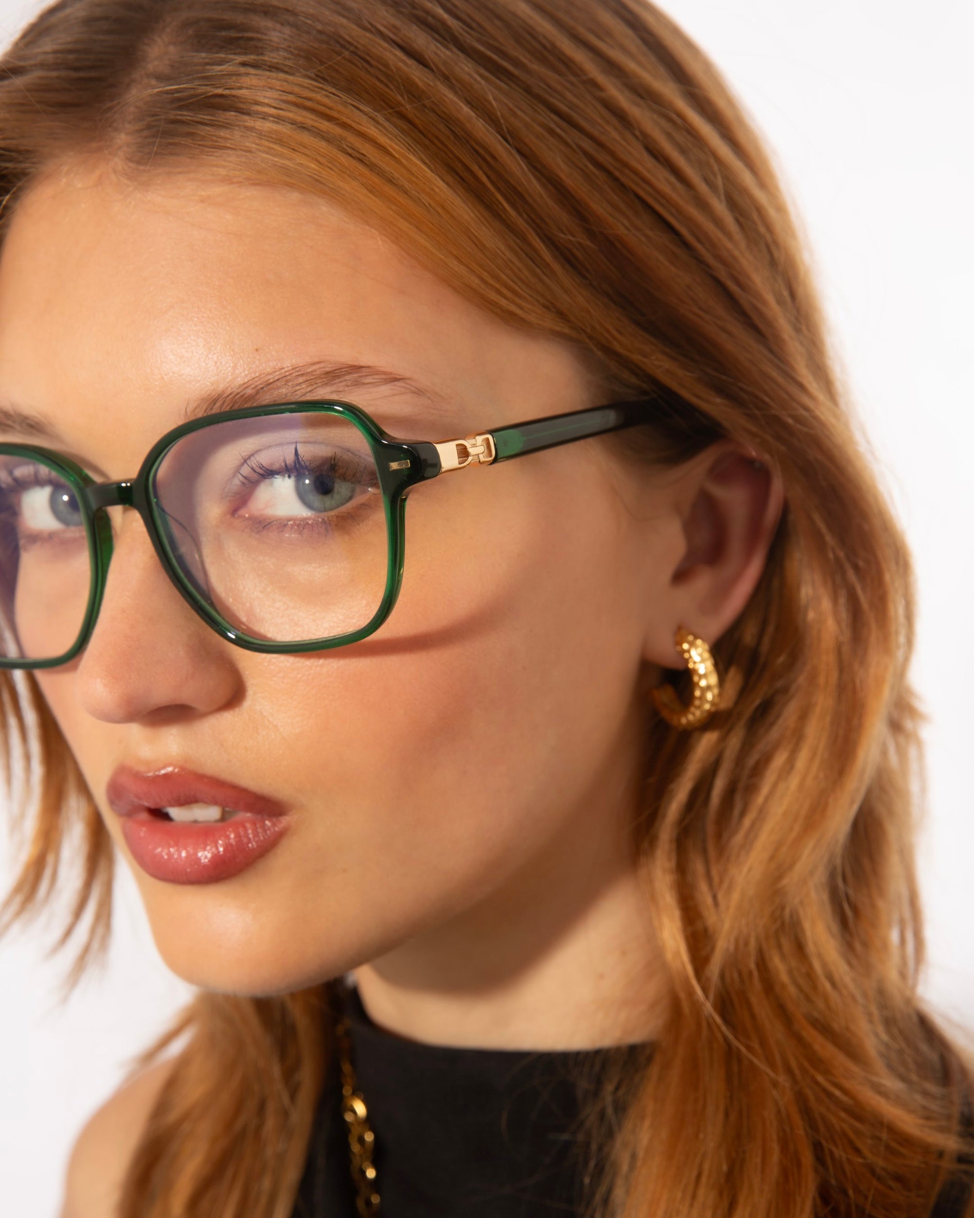 A person with long, light brown hair wears large, green-framed For Art's Sake® Charm cat-eye shaped glasses and hoop earrings. They have a neutral expression and are looking at the camera. The background is plain white.