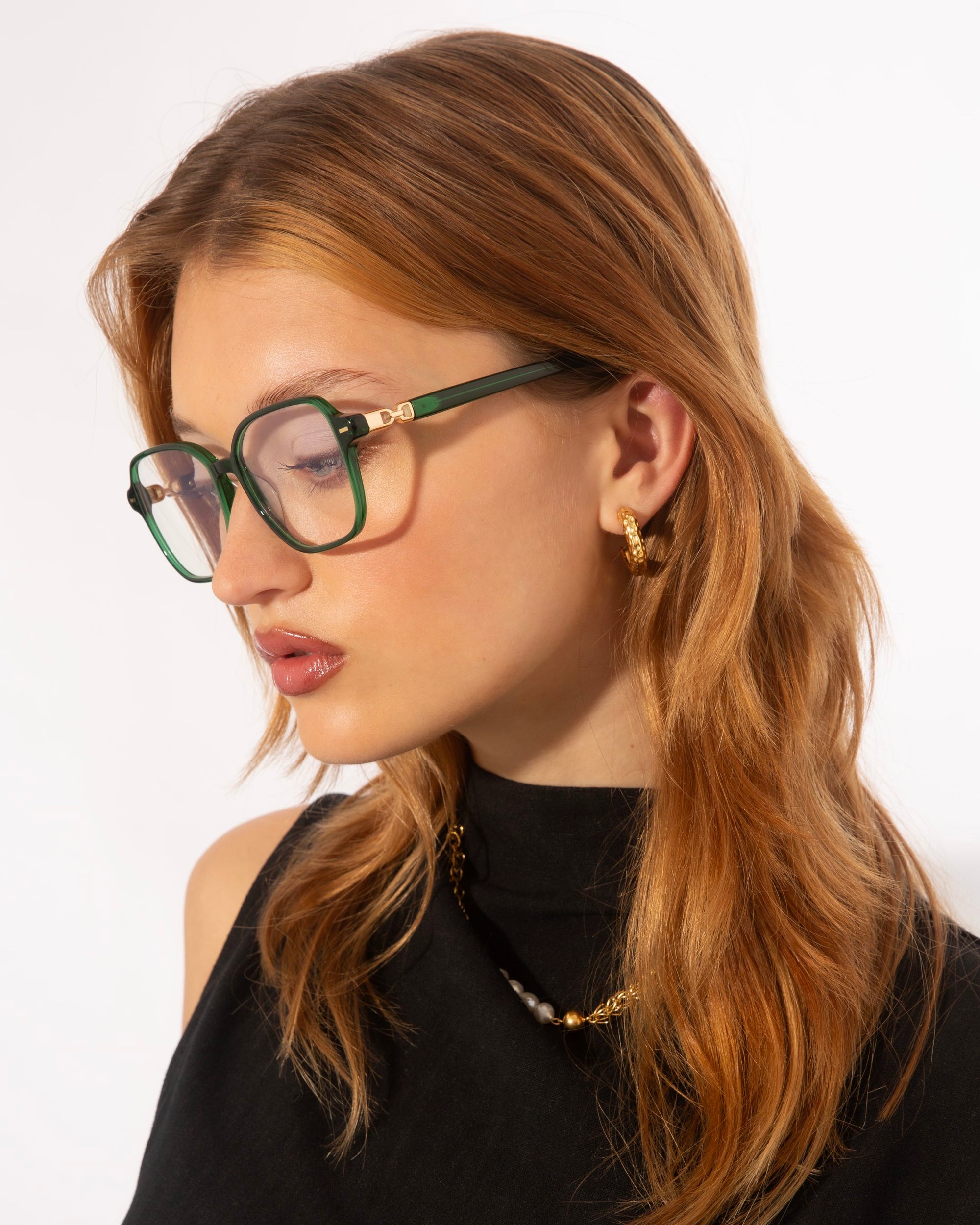 A woman with long, red hair is shown in a profile view against a plain background. She is wearing green-framed Charm glasses by For Art's Sake®, gold hoop earrings, and a black sleeveless top. Her serious expression is accentuated by her slightly parted lips.