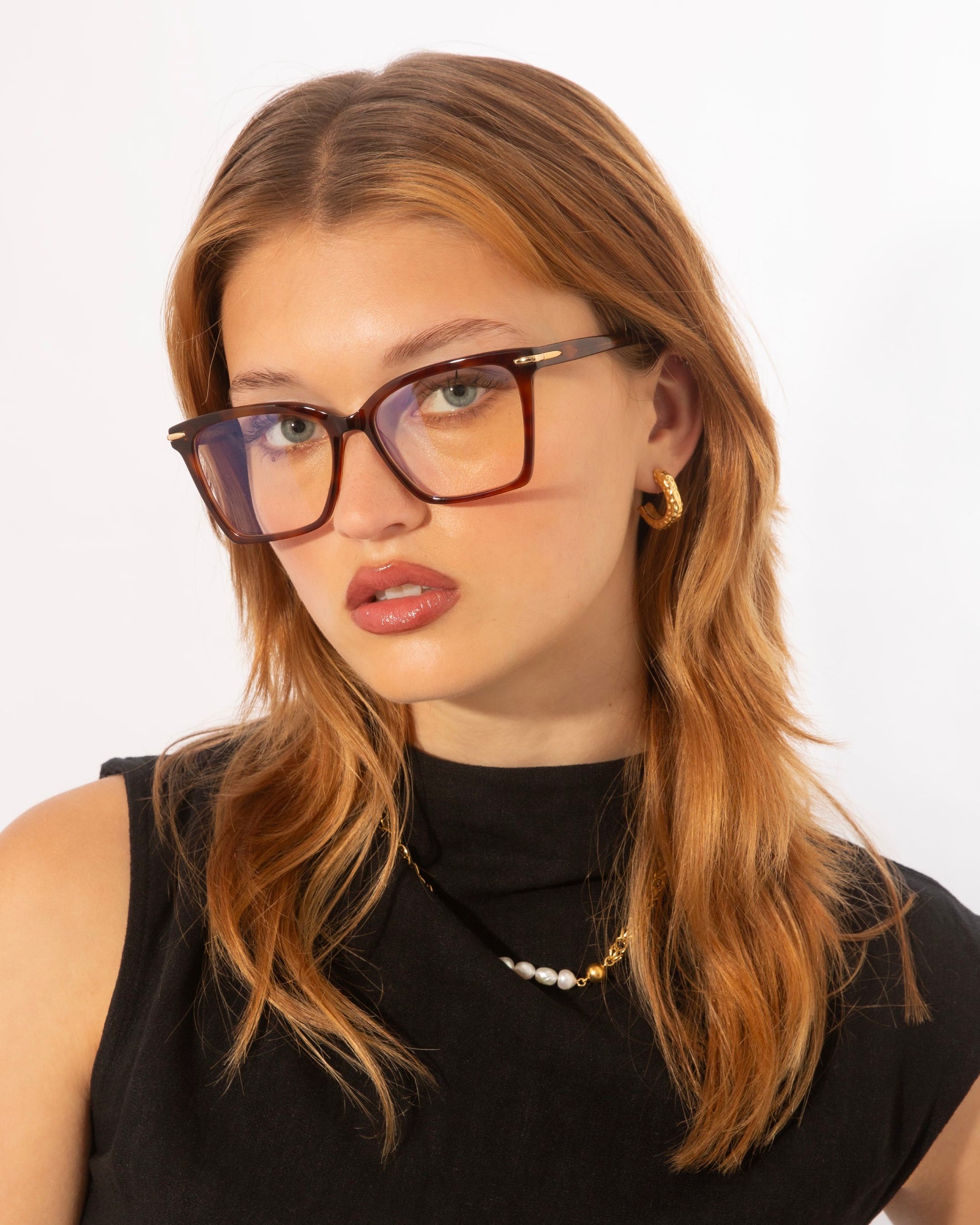 A woman with long, light brown hair is wearing rectangular Azure optical glasses by For Art&#39;s Sake® with a brown frame. She has a serious expression, with her head slightly tilted. Dressed in a sleeveless black top and accessorized with small hoop earrings and a delicate necklace, she stands against a white background.