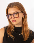 A woman with long, light brown hair is wearing rectangular Azure optical glasses by For Art's Sake® with a brown frame. She has a serious expression, with her head slightly tilted. Dressed in a sleeveless black top and accessorized with small hoop earrings and a delicate necklace, she stands against a white background.