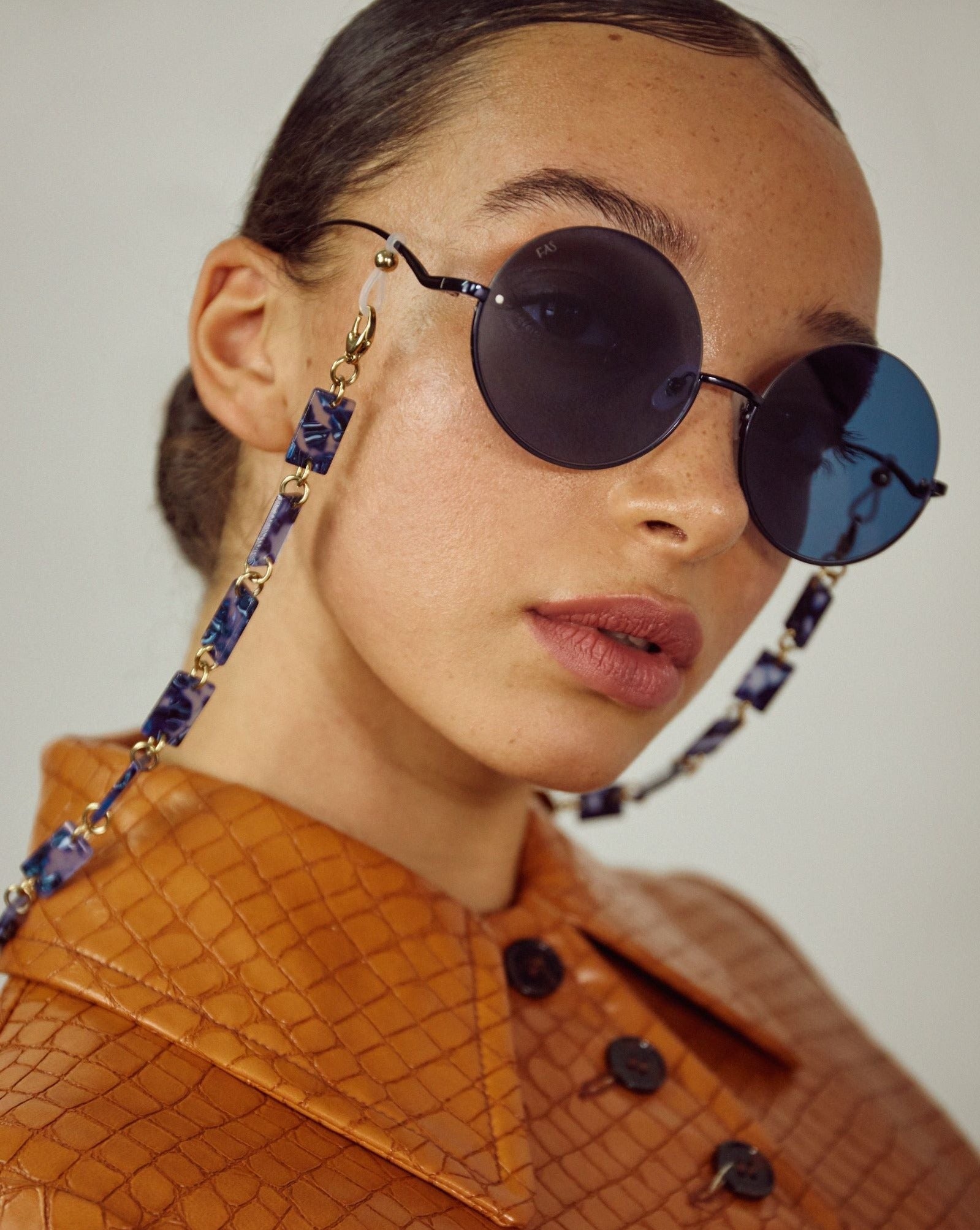 A person with slicked-back hair is wearing round, dark sunglasses with stainless steel frames and a decorative chain. They are dressed in a brown, textured jacket with a buttoned front. The background is plain and off-white. The sunglasses they are wearing are the Oceana by For Art&#39;s Sake®.
