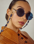 A person with slicked-back hair is wearing round, dark sunglasses with stainless steel frames and a decorative chain. They are dressed in a brown, textured jacket with a buttoned front. The background is plain and off-white. The sunglasses they are wearing are the Oceana by For Art's Sake®.