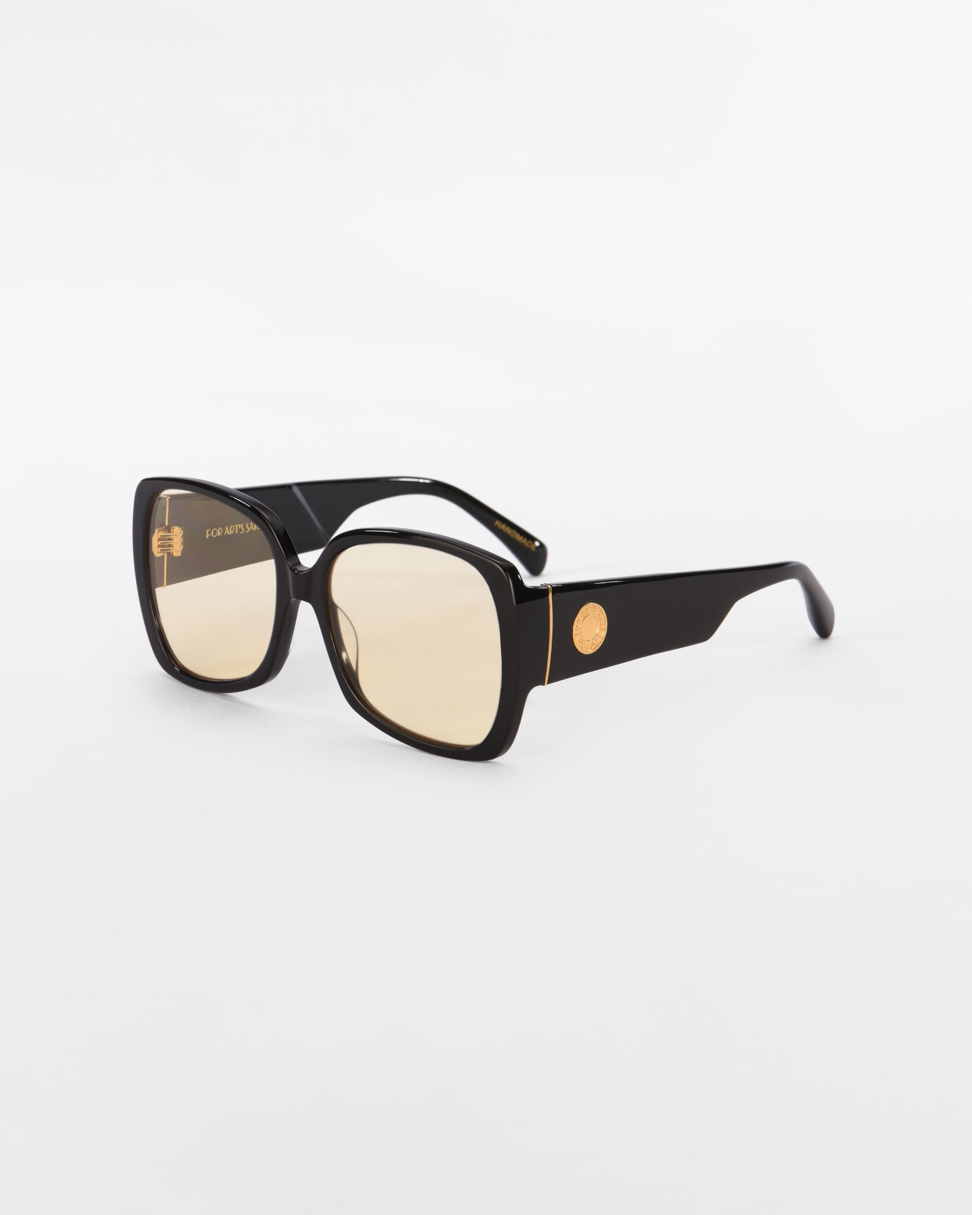 A pair of stylish, oversized black For Art&#39;s Sake® Odyssey sunglasses with rectangular lenses and gold accents on the temples, including a small emblem. The lightweight nylon lenses have a slight amber tint, complemented by 18-karat gold plating, and the background is plain white.