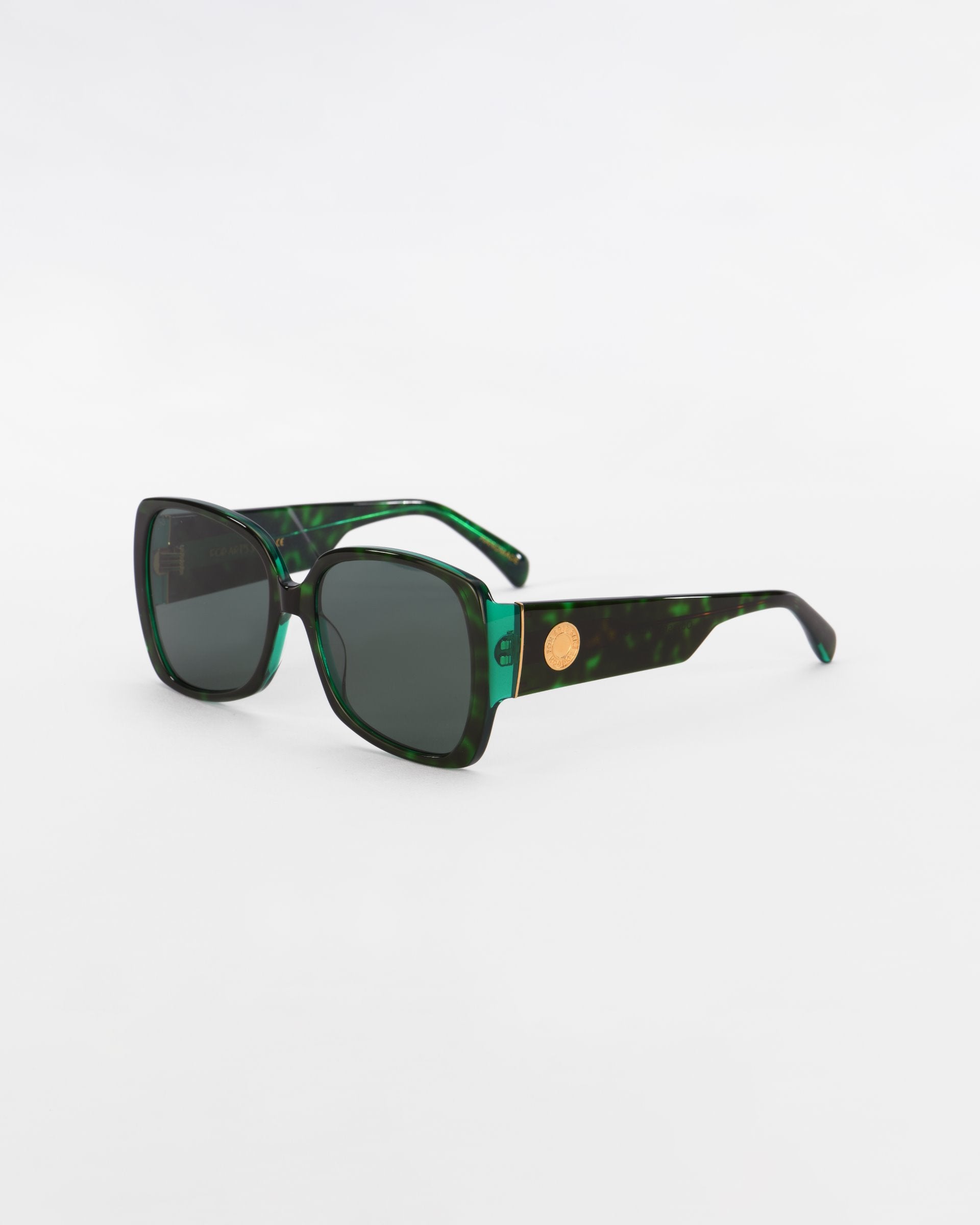 A pair of dark green tortoiseshell pattern Odyssey sunglasses by For Art&#39;s Sake® with large, square frames and lightweight nylon lenses. The temples are thick and feature a small circular emblem with 18-karat gold plating near the hinges. The background is plain white.
