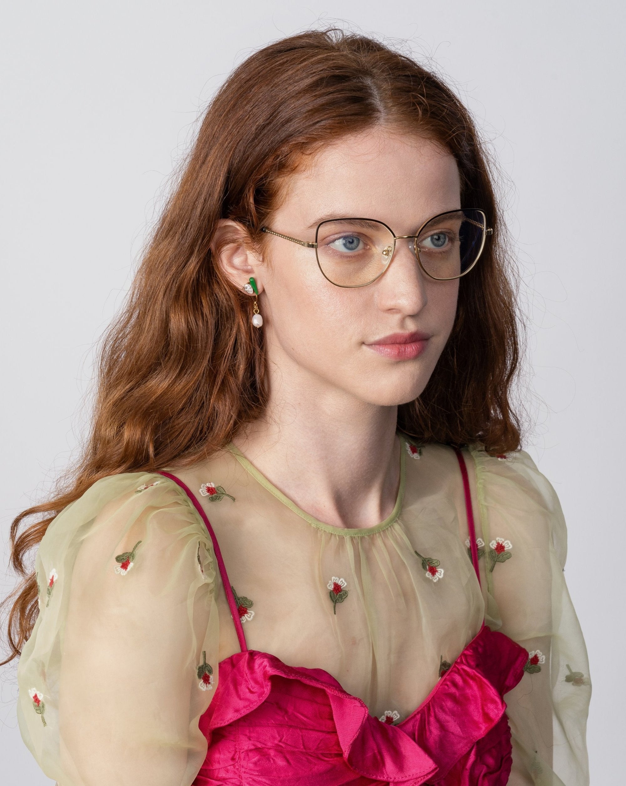 A woman with long, wavy red hair, wearing Ophelia glasses by For Art&#39;s Sake® with a cat-eye silhouette and green-and-gold earrings, is dressed in a sheer, light green blouse with embroidered flowers over a bright pink, textured top. She is looking slightly to the side against a plain, light gray background.