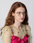 A woman with long, wavy red hair, wearing Ophelia glasses by For Art's Sake® with a cat-eye silhouette and green-and-gold earrings, is dressed in a sheer, light green blouse with embroidered flowers over a bright pink, textured top. She is looking slightly to the side against a plain, light gray background.