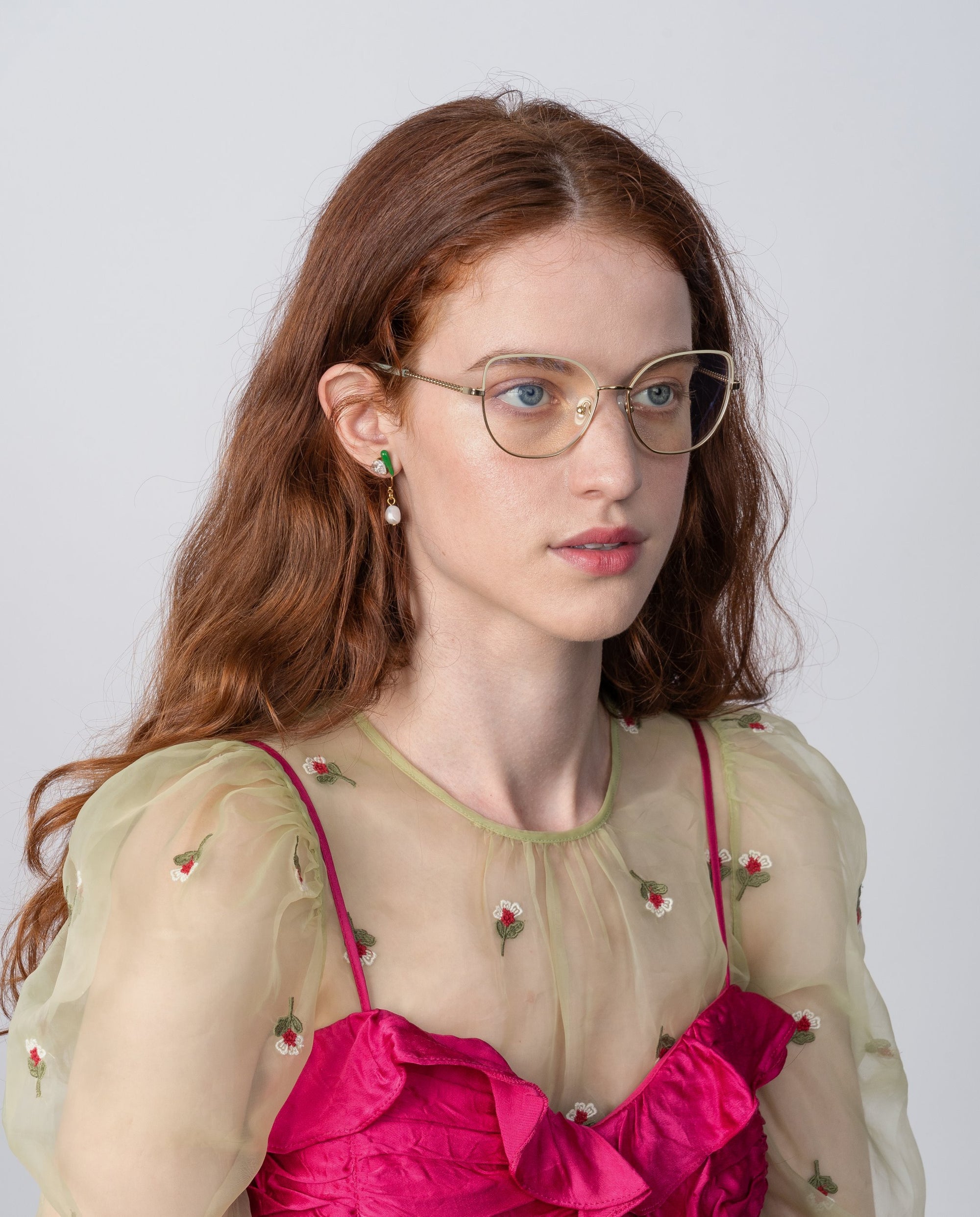 A woman with long, wavy auburn hair and wearing Ophelia glasses by For Art's Sake® is looking slightly to the side. She is dressed in a sheer, green, floral-embroidered top over a bright pink, ruffled dress. The background is plain and light-colored.