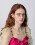 A woman with long, wavy auburn hair and wearing Ophelia glasses by For Art's Sake® is looking slightly to the side. She is dressed in a sheer, green, floral-embroidered top over a bright pink, ruffled dress. The background is plain and light-colored.