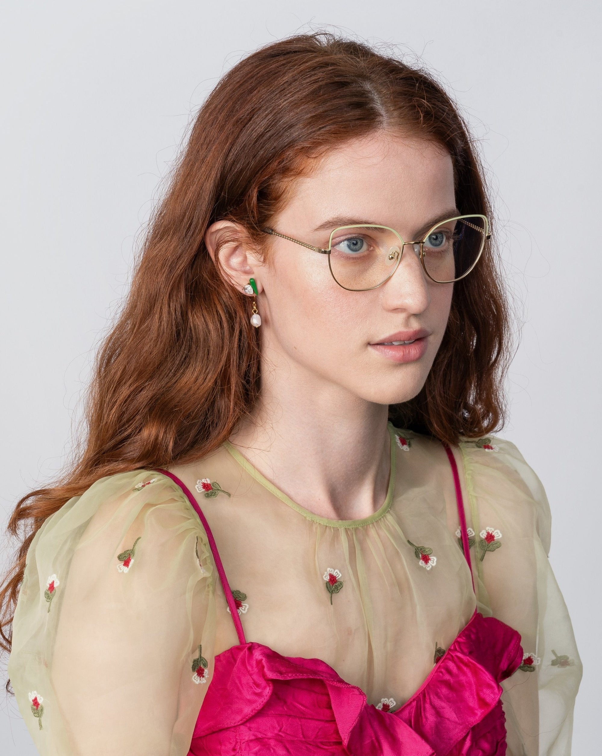 A woman with long, wavy red hair is wearing Ophelia glasses by For Art&#39;s Sake® with a cat-eye silhouette and a sheer beige top adorned with small embroidered flowers over a bright pink undergarment. She has green and pink earrings, and she is looking slightly to the side against a plain, light gray background.