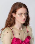 A woman with long, wavy red hair is wearing Ophelia glasses by For Art's Sake® with a cat-eye silhouette and a sheer beige top adorned with small embroidered flowers over a bright pink undergarment. She has green and pink earrings, and she is looking slightly to the side against a plain, light gray background.