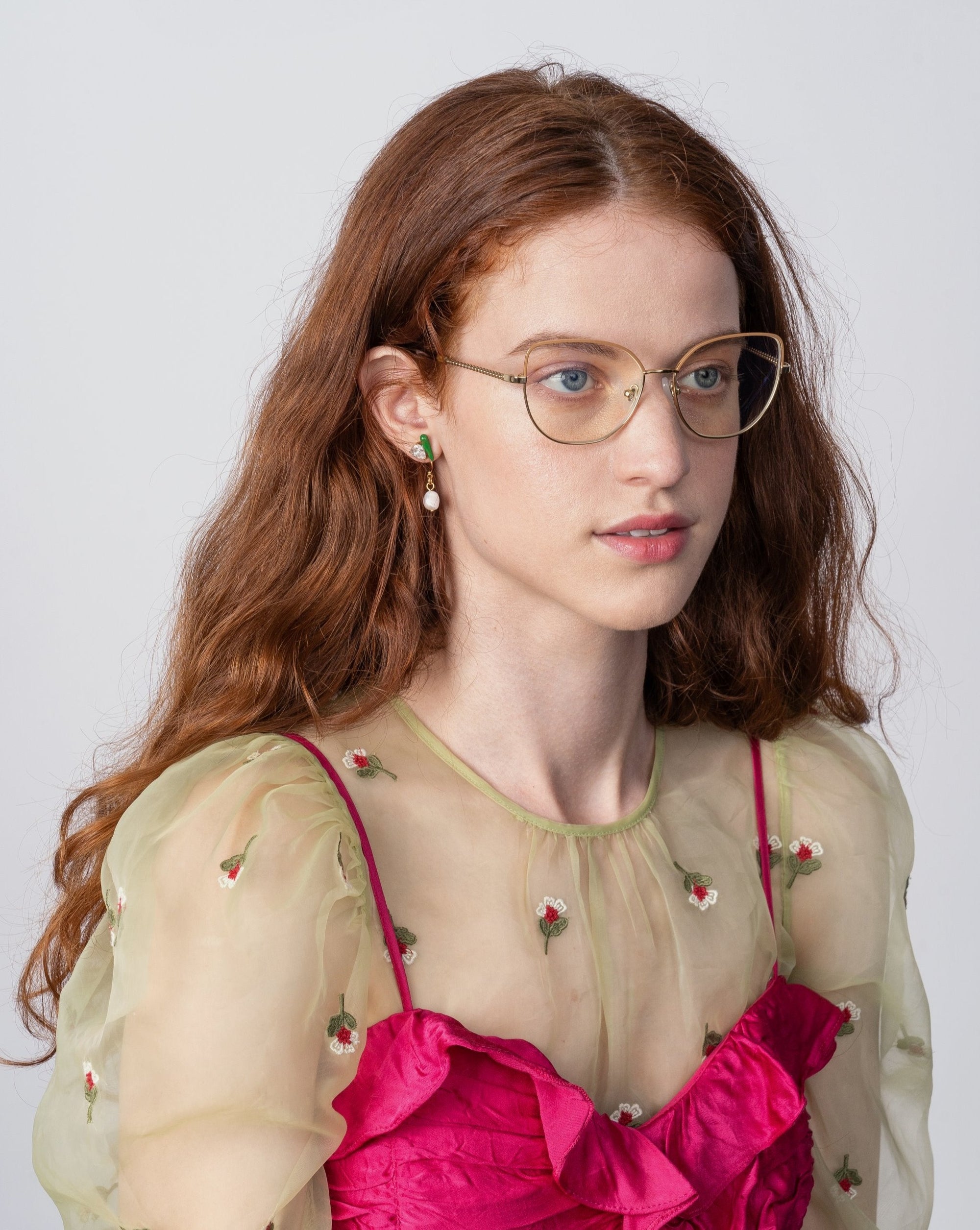 A woman with long, wavy red hair and glasses with a cat-eye silhouette is wearing a sheer green top adorned with small floral designs over a bright pink dress. She has a calm expression and is looking slightly to the side, wearing the Ophelia by For Art&#39;s Sake®. The background is plain and light-colored.