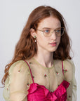 A woman with long, wavy red hair and glasses with a cat-eye silhouette is wearing a sheer green top adorned with small floral designs over a bright pink dress. She has a calm expression and is looking slightly to the side, wearing the Ophelia by For Art's Sake®. The background is plain and light-colored.