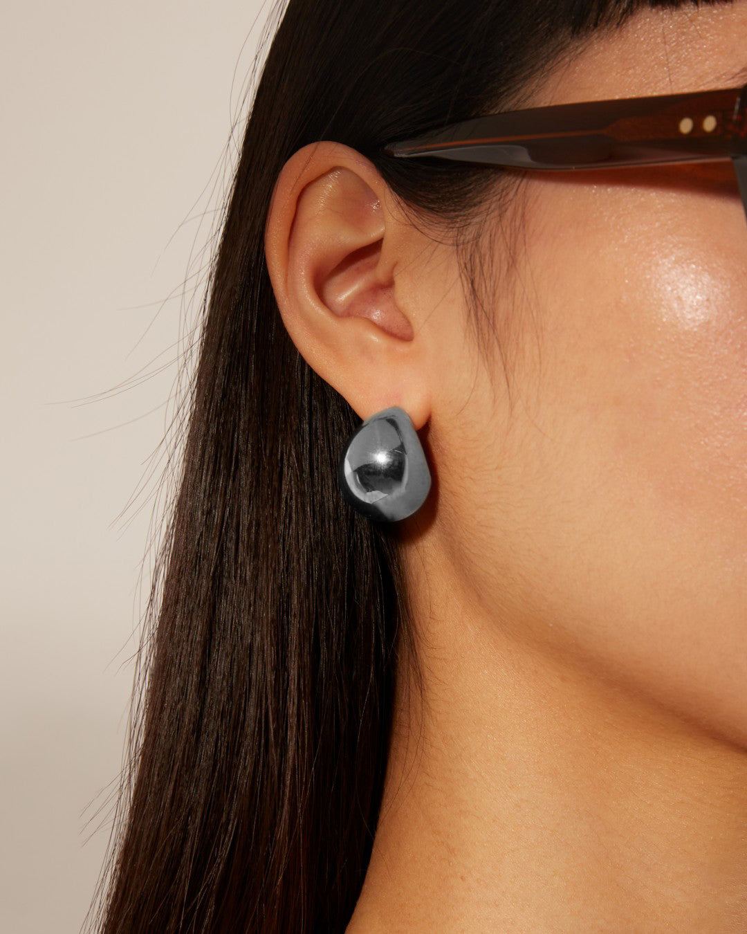 A close-up image of a person with long, dark hair wearing shiny, teardrop-shaped For Art's Sake® Orbit Earrings Silver. The individual is also wearing large, dark-framed glasses. The background is a neutral, light color.