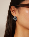 A close-up image of a person with long, dark hair wearing shiny, teardrop-shaped For Art's Sake® Orbit Earrings Silver. The individual is also wearing large, dark-framed glasses. The background is a neutral, light color.