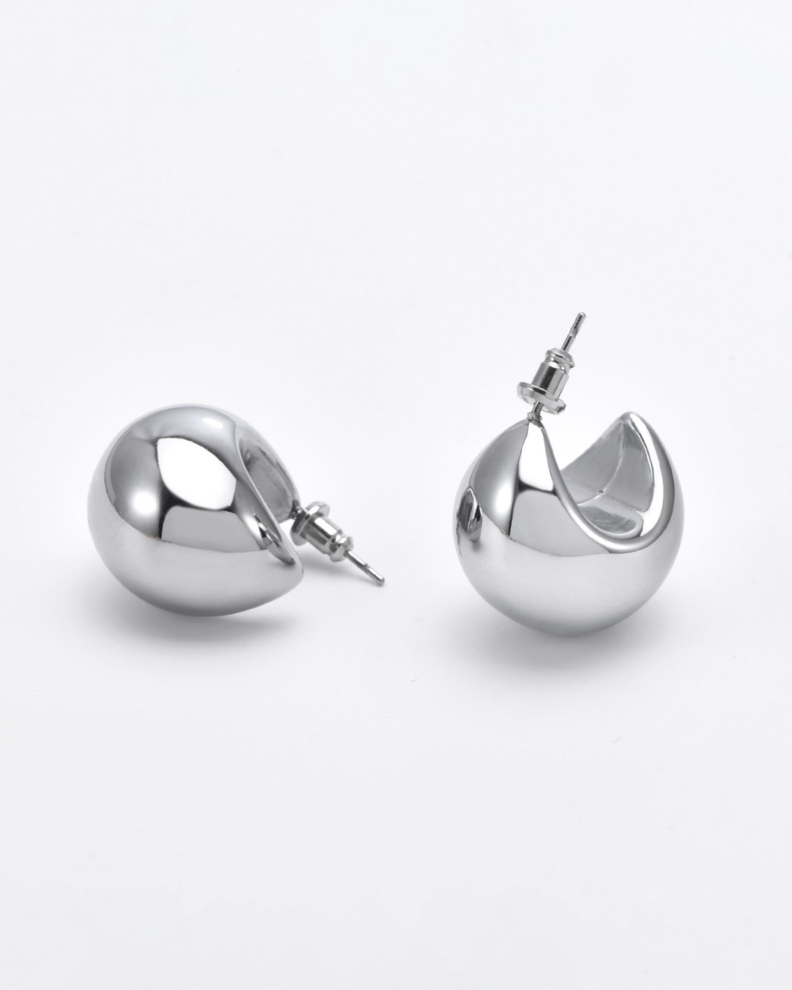 Close-up of a pair of shiny, hypoallergenic Orbit Earrings Silver by For Art's Sake®. One earring is lying on its side, showing a smooth, rounded surface. The other is upright, revealing an open, curved back reminiscent of For Art's Sake® earrings. The background is a plain white surface.