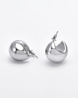 Close-up of a pair of shiny, hypoallergenic Orbit Earrings Silver by For Art's Sake®. One earring is lying on its side, showing a smooth, rounded surface. The other is upright, revealing an open, curved back reminiscent of For Art's Sake® earrings. The background is a plain white surface.