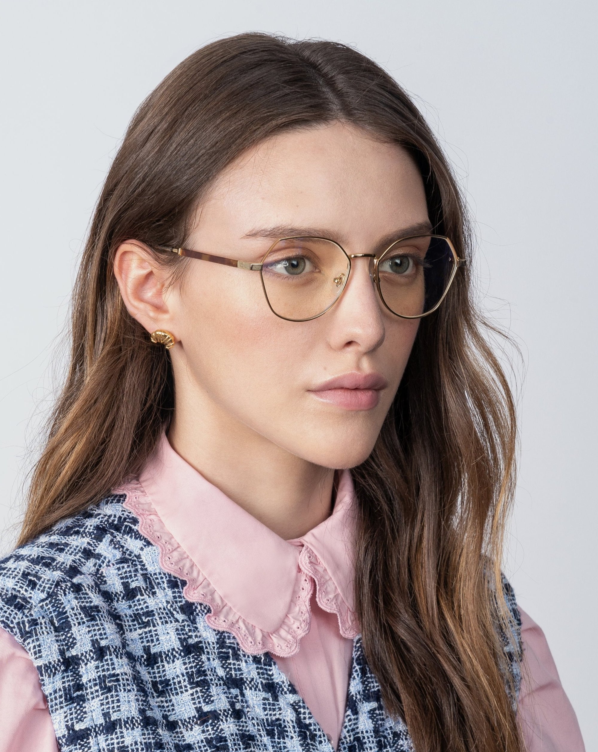 A woman with long, brown hair wearing 18-karat gold-plated glasses and gold earrings. She is dressed in a pink blouse with a lace collar under a blue and white checkered vest. Her glasses have Orchard blue light filter prescription lenses by For Art's Sake®. She has a calm, neutral expression and is facing slightly to the right.