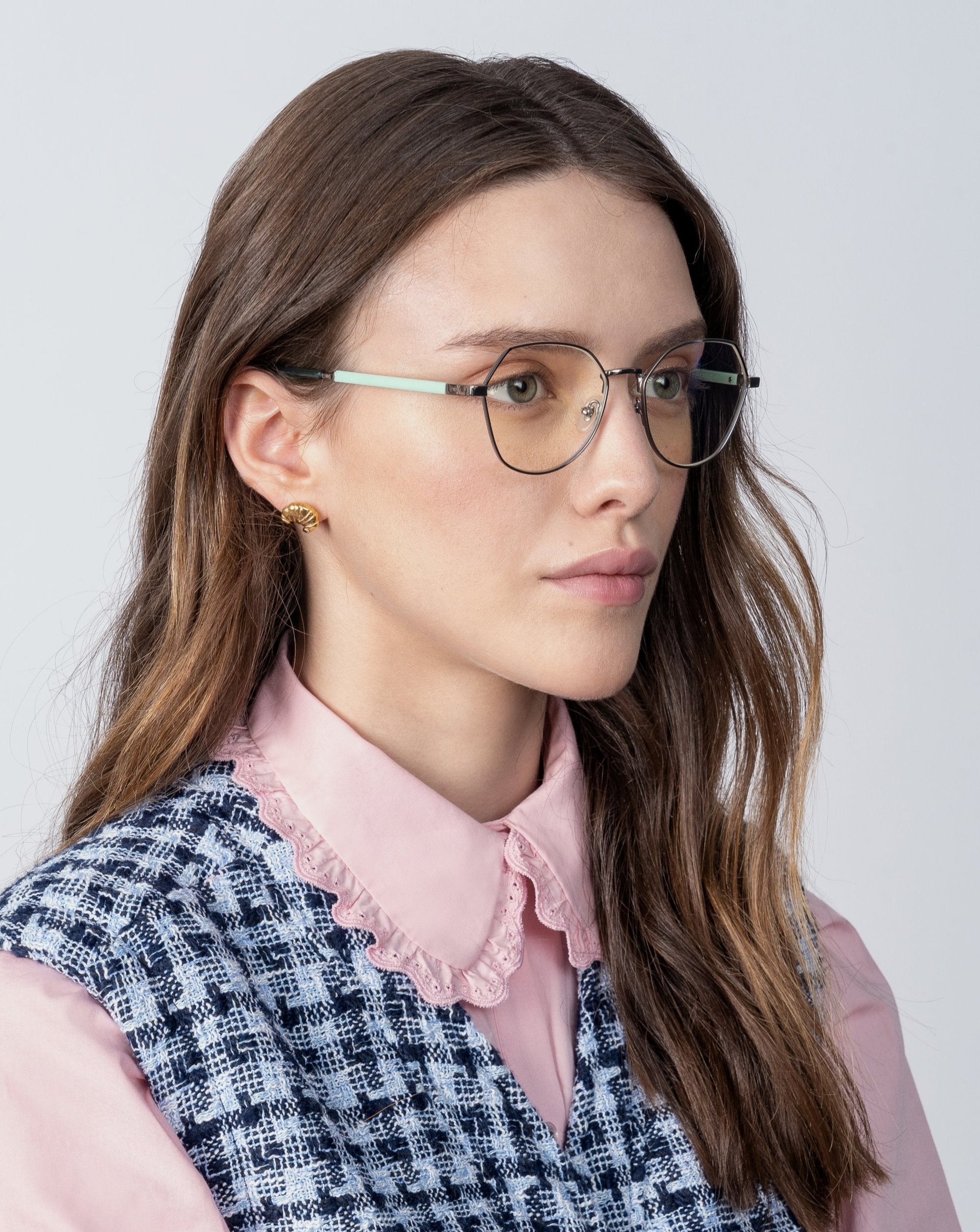 A young woman with long, wavy brown hair is wearing round, prescription Orchard lenses and metal-framed glasses by For Art's Sake®. She is dressed in a pink collared shirt with a lace trim and a blue patterned vest. She faces slightly to the left against a plain light background.