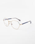 Close-up of a pair of Orchard frames by For Art's Sake® with clear prescription lenses, featuring an angular design and navy-blue accents on the tips of the arms. The glasses are positioned against a plain white background.