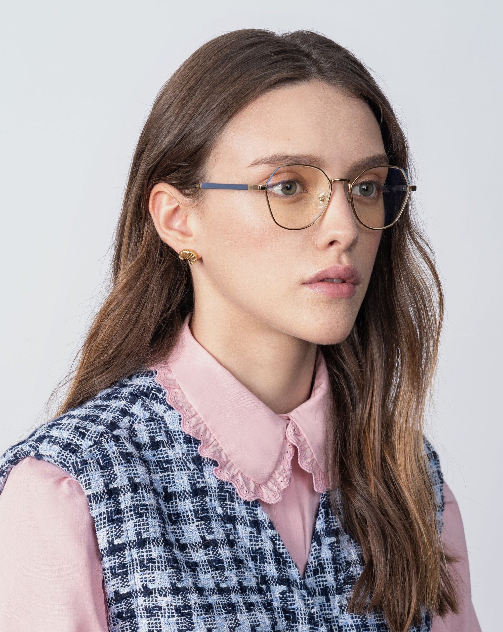 A woman with long hair wearing gold-rimmed For Art&#39;s Sake® Orchard glasses featuring prescription lenses and a blue light filter, a pink blouse with a lace collar, and a blue and white plaid vest. She has small 18-karat gold-plated earrings and is looking slightly to the left with a neutral expression. The background is plain and light-colored.