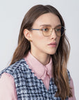 A woman with long hair wearing gold-rimmed For Art's Sake® Orchard glasses featuring prescription lenses and a blue light filter, a pink blouse with a lace collar, and a blue and white plaid vest. She has small 18-karat gold-plated earrings and is looking slightly to the left with a neutral expression. The background is plain and light-colored.