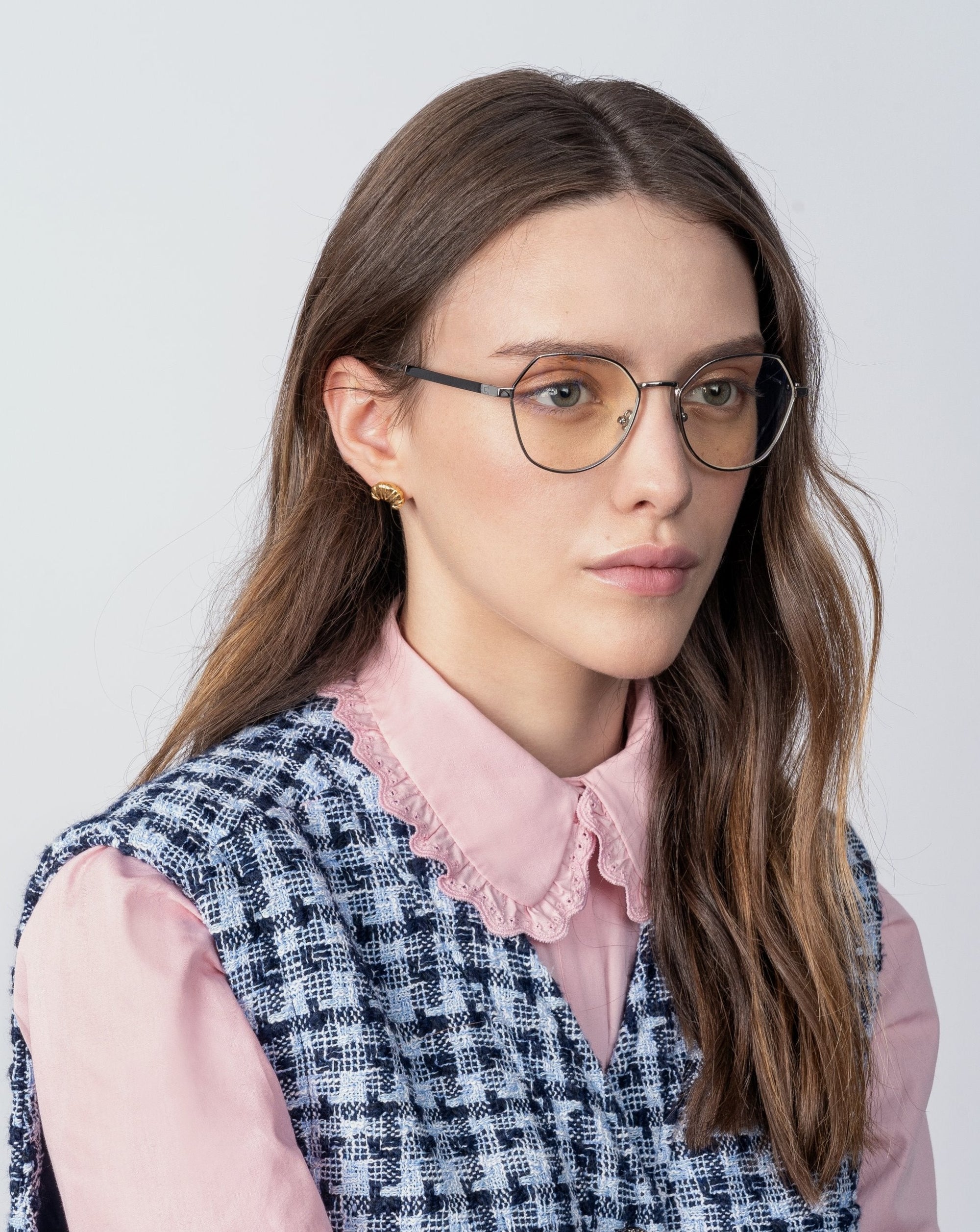 A woman with long brown hair wears Orchard round glasses with gold-plated frames by For Art&#39;s Sake®, a pink blouse with a lace collar, and a blue and white checkered vest. She has a neutral expression and is looking slightly to the side. The background is plain light gray.