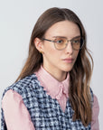 A woman with long brown hair wears Orchard round glasses with gold-plated frames by For Art's Sake®, a pink blouse with a lace collar, and a blue and white checkered vest. She has a neutral expression and is looking slightly to the side. The background is plain light gray.