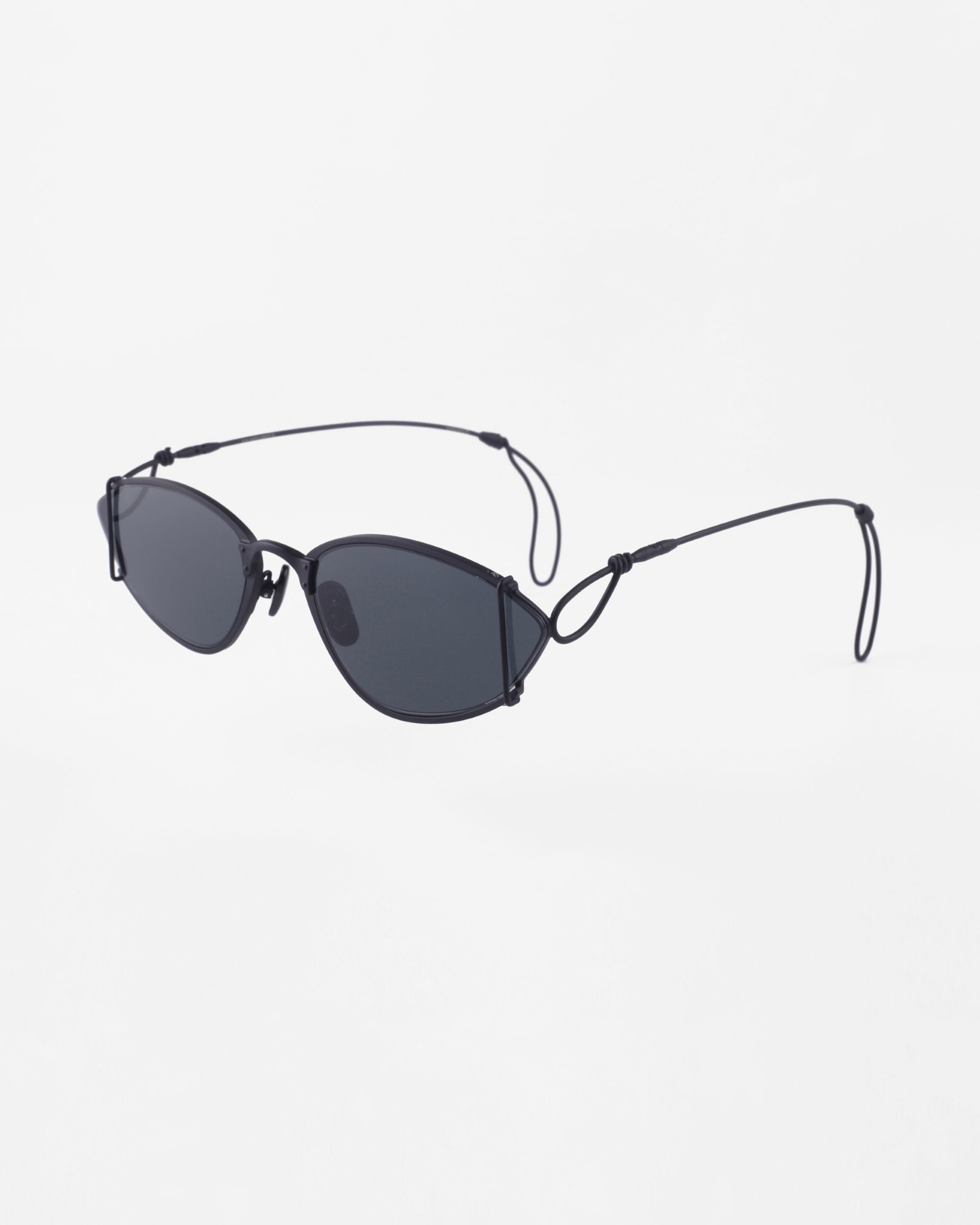A pair of sleek, black, oval-shaped For Art&#39;s Sake® Ornate sunglasses with thin wire arms and adjustable earpieces. The minimalist design features dark lenses with 100% UVA &amp; UVB protection and a lightweight frame positioned against a white background.