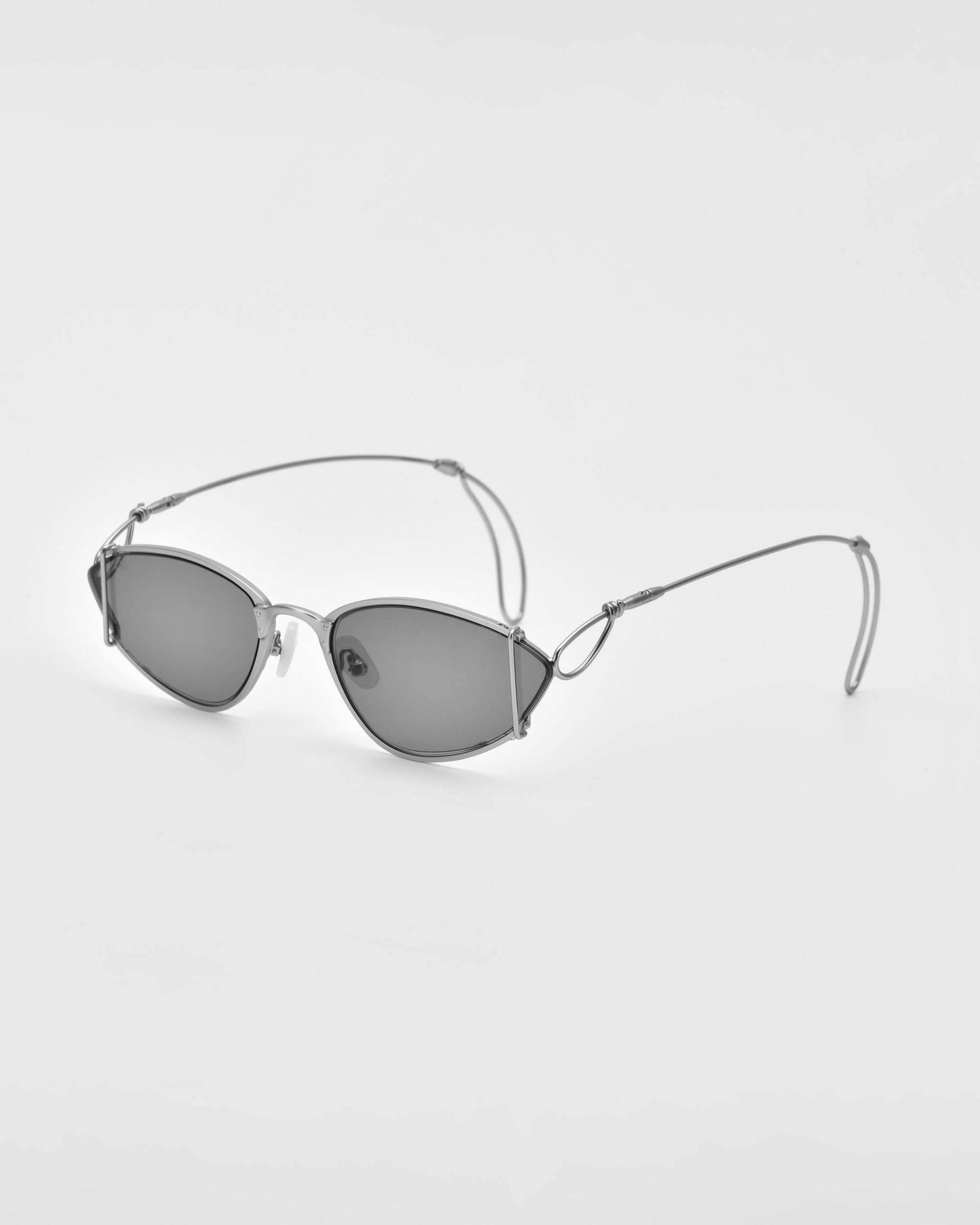 A pair of Ornate sunglasses by For Art&#39;s Sake® with sleek, thin metal frames and oval dark lenses. The arms feature intricate wire details, giving them a modern minimalist look while providing essential UV protection. The background is plain white.