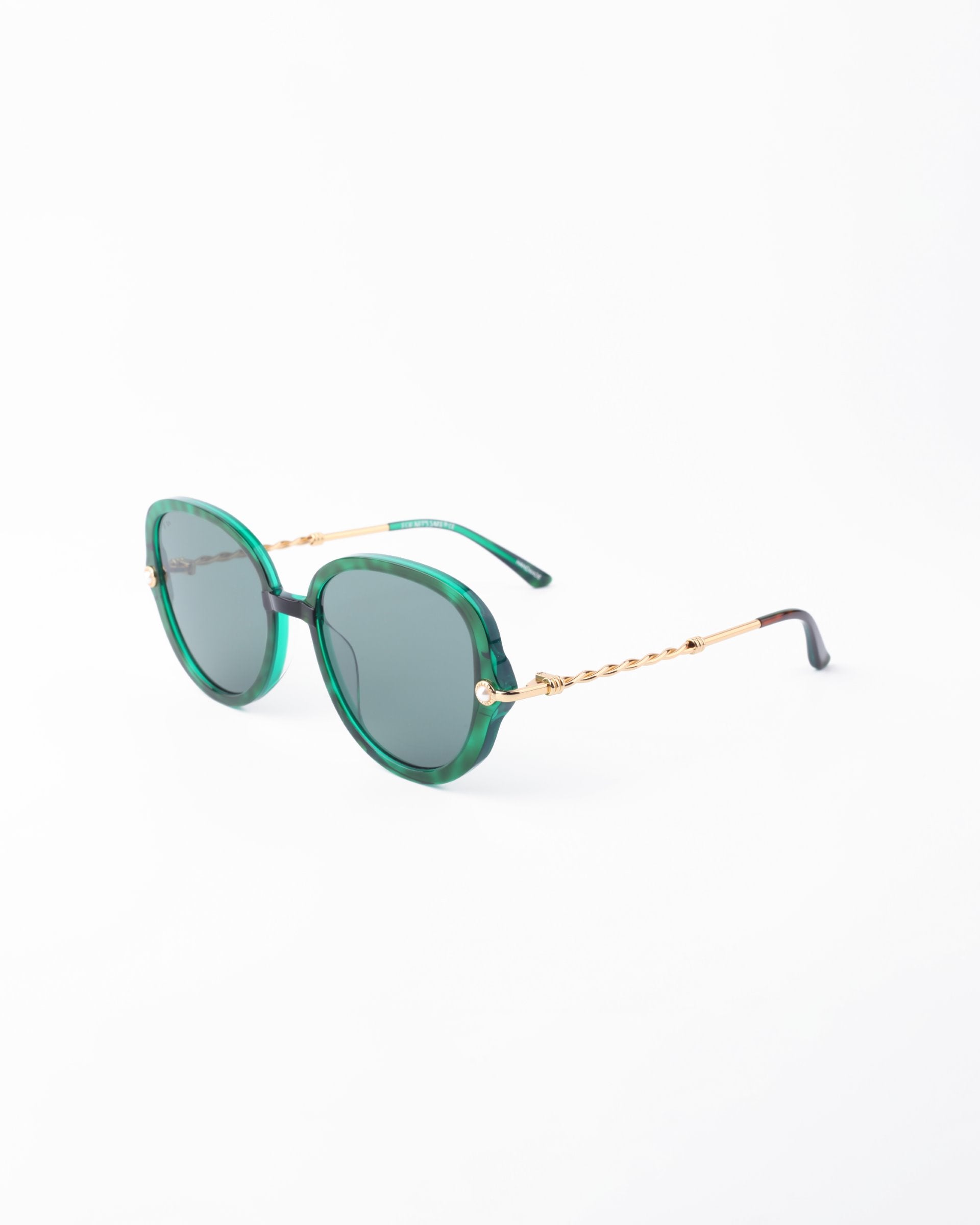 A pair of stylish For Art&#39;s Sake® Primrose sunglasses with green-tinted round lenses and handmade gold-plated metal arms. The frames are green with a glossy finish, made from plant-based acetate, and the twisted design on the arms adds a unique touch. The ends of the arms are black, and they feature an anti-reflective coating for enhanced comfort. Displayed on a white background.