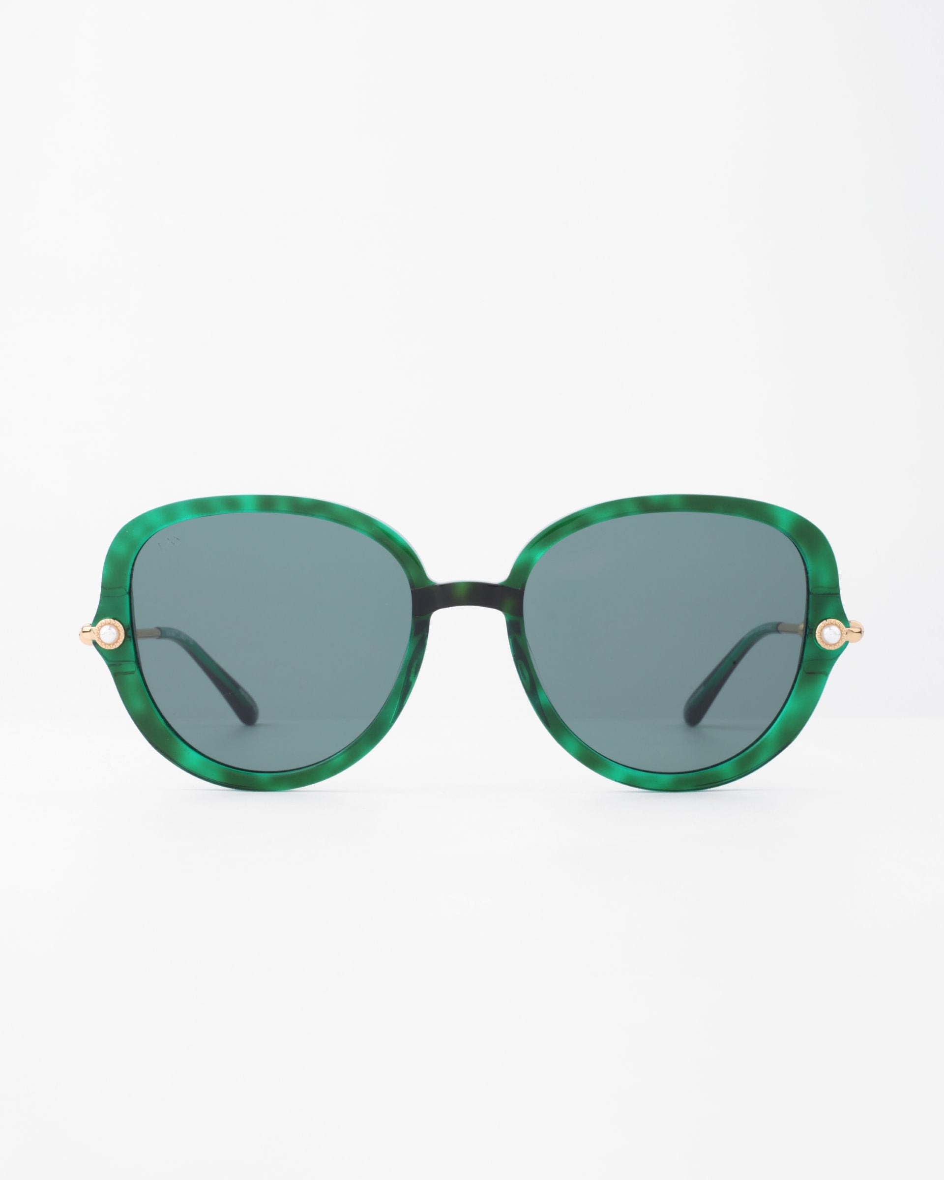 A pair of stylish green round sunglasses with dark tinted lenses are centered against a plain white background. The Primrose sunglasses by For Art&#39;s Sake® have gold accents on the hinges, adding a touch of elegance, and feature a plant-based acetate frame for an eco-friendly twist.