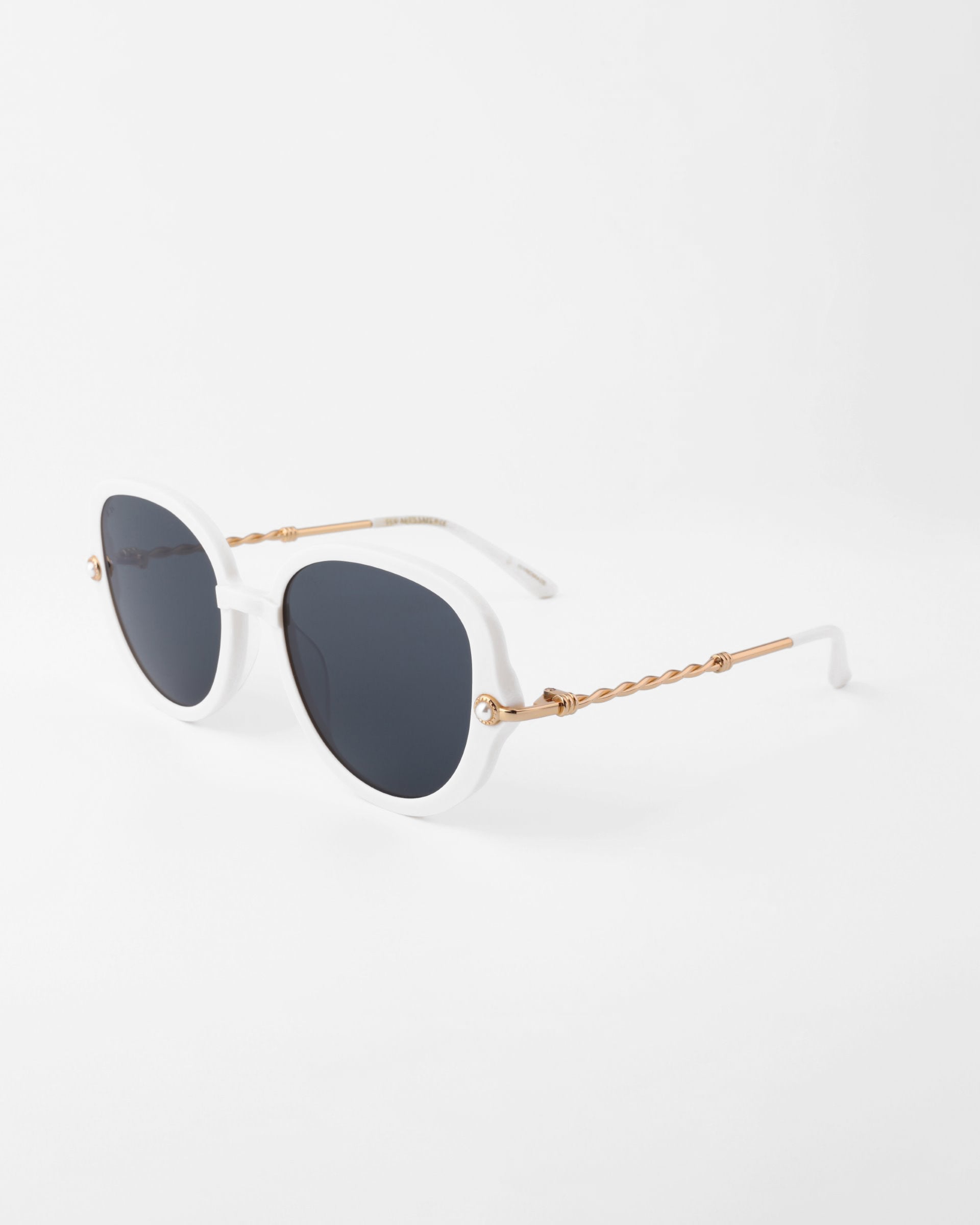 A pair of stylish round sunglasses featuring plant-based acetate white frames, dark lenses, and intricate handmade gold-plated temples with a twisted design, placed against a plain white background. The Primrose by For Art&#39;s Sake®.
