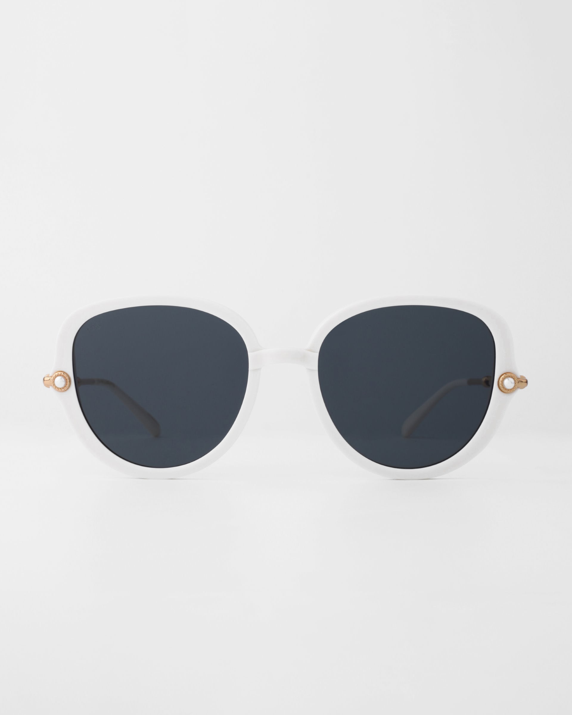 A pair of white-framed For Art's Sake® Primrose sunglasses with large, round dark lenses. Each temple is adorned with a small, handmade gold-plated decorative detail near the hinges. The glossy, smooth frame is made from plant-based acetate and the background is plain white.