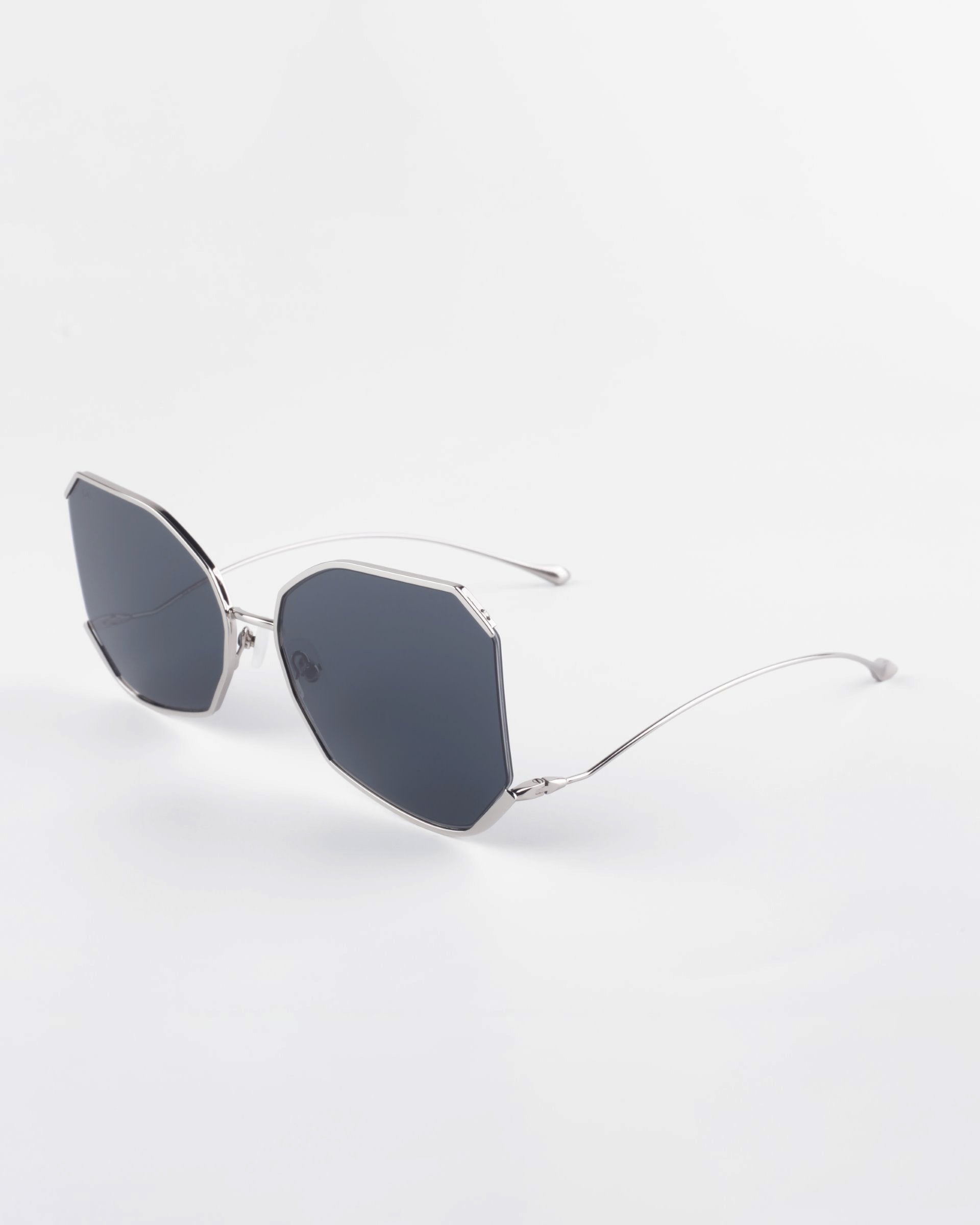 A pair of modern, silver-framed sunglasses with unique angular, dark-tinted lenses is displayed against a plain white background. The gold-plated stainless steel design features sleek, thin arms adding to the minimalist aesthetic and 100% UVA &amp; UVB protection. The product name is Painter from For Art&#39;s Sake®.