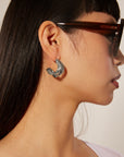 A person wearing large, dark sunglasses and a distinctive, silver, wave-shaped earring that curls around the earlobe. The person has long, straight hair, adorned with a palladium-plated hoop earring as well. They are shown in profile against a neutral background wearing Portrait Earrings Silver by For Art's Sake®.