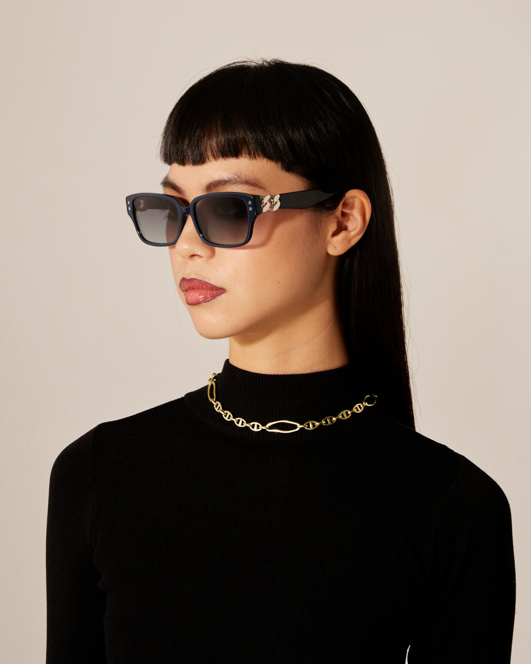 A person with straight black hair and bangs is wearing rectangular sunglasses with dark lenses, a black turtleneck, and a Portrait Necklace Gold by For Art&#39;s Sake®. The background is neutral.