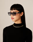A person with straight black hair and bangs is wearing rectangular sunglasses with dark lenses, a black turtleneck, and a Portrait Necklace Gold by For Art's Sake®. The background is neutral.