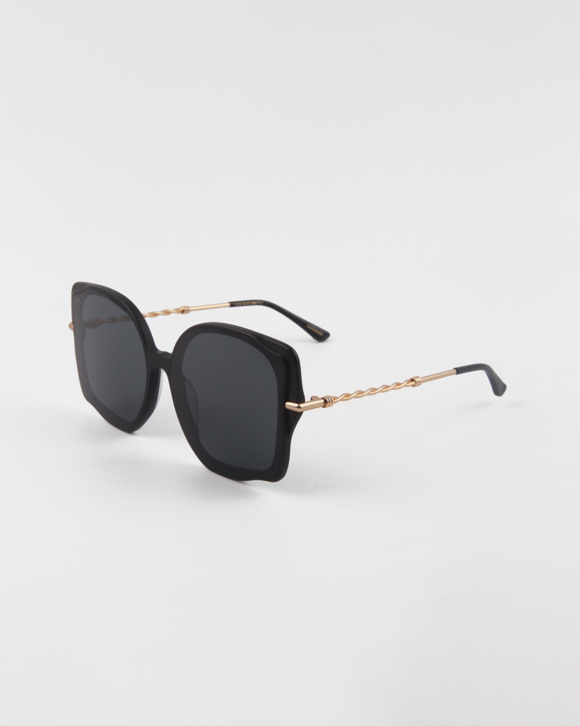 A pair of chic, black square-shaped Fahrenheit handmade acetate sunglasses by For Art's Sake® with dark lenses. The oversized frames have 18-karat gold-plated arms featuring a twisted design near the hinges. The sunglasses rest on a white surface with a gentle shadow.