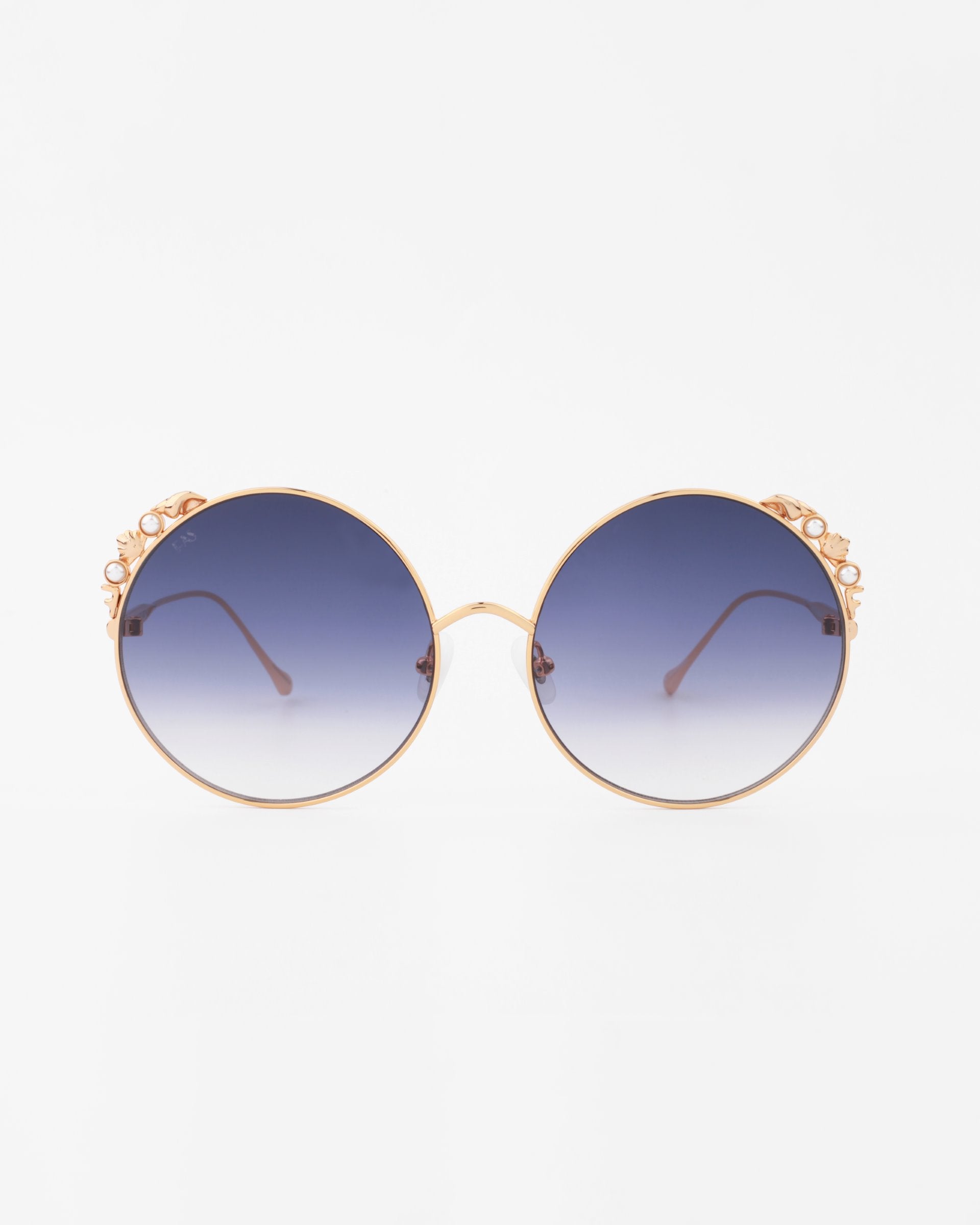 A pair of For Art's Sake® Lindy handmade gold-plated round sunglasses with ultra-lightweight shatter-resistant dark blue gradient lenses. The bridge and end pieces have a decorative floral embellishment, offering 100% UVA & UVB protection. The For Art's Sake® Lindy sunglasses are set against a plain white background.